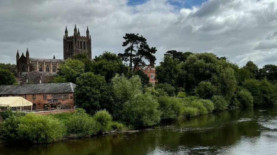 A flowing river with trees and bushes alongside, historical buildings and a cathedral in the background.