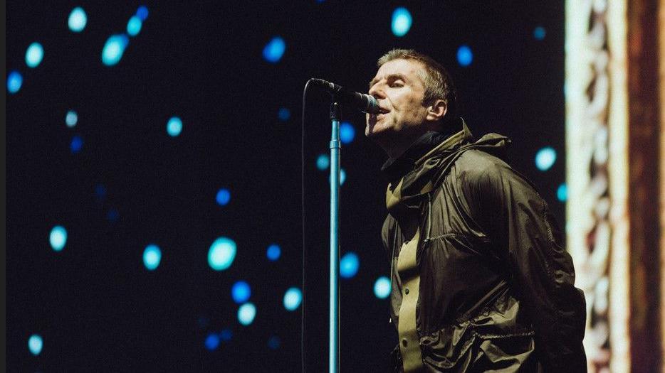 Liam Gallagher is singing, with light and darker blue circles of light behind him.