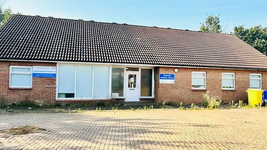 Exterior of the GP surgery- a bring building with a a white glass door and windows and overgrown weeds in front of it 