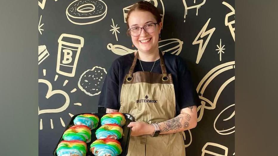 Chloe Morrison standing in front of a black background featuring drawings of coffee cups, hearts and donuts. She is wearing a beige Butterwick apron whilst holding a tray of rainbow-coloured donuts.