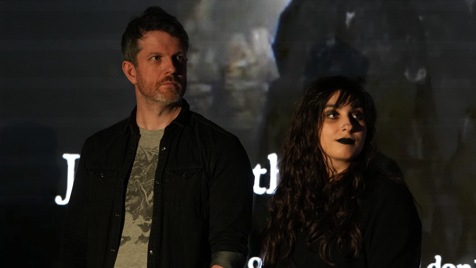 Two people stand in front of a screen, a man on the left and a woman on the right. The man wears a black jacket with a grey t-shirt. He has grey hair. The woman has long black hair and wears a black top.