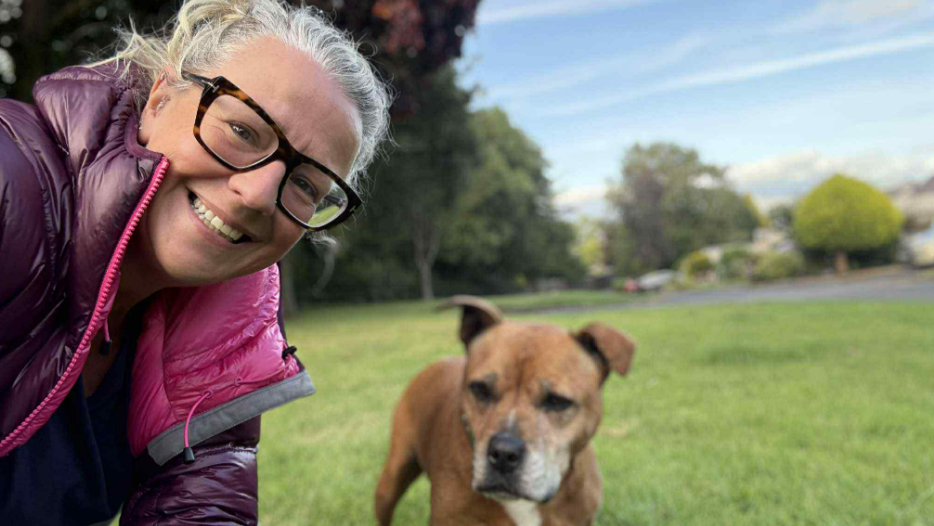 Ellie smiling into the camera, crouched next to a brown, terrier dog in a green field. She has her hair tied back and is wearing large brown glasses. She is also wearing a purple puffer jacket.