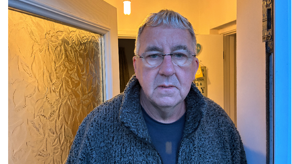 David standing outside his front door with a frustrated look. He wears a grey t-shirt, a grey fleece and clear glasses. David has short grey hair.