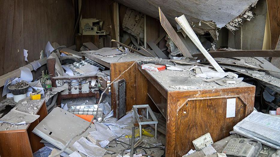 Damaged office equipment sits among the destruction at Evin Prison after it was hit by an Israeli air strikes. The ceiling is collapsed and piles of paperwork are strewn about. A wooden desk is in the centre of the room.