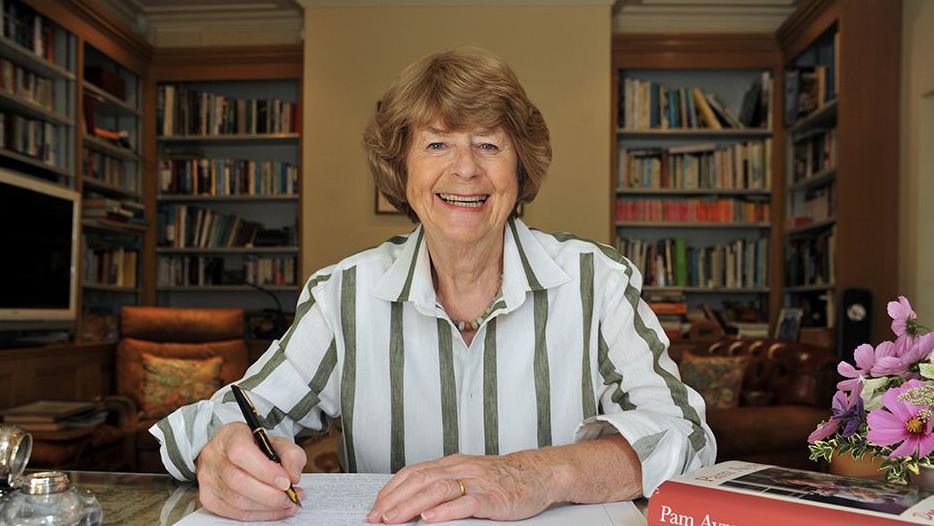 Pam Ayres smiling and sitting at a desk in an office in a home writing as she writes in a book with a fountain pen in her hand and one of her books on the right of her desk