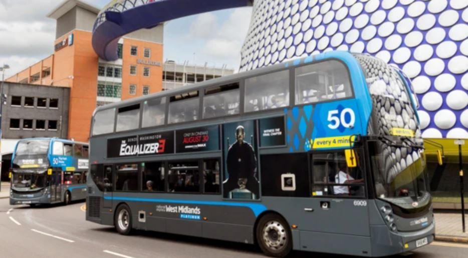 A grey double decker bus on a city street. The bus has a poster for the Equalizer 3 on the side and the number "50" in white letters. It is driving past a purple building with lots of white circles on it, with another bus driving behind it.