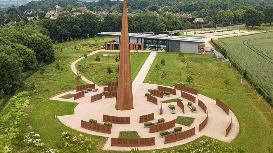 A copper-coloured metal spire in the middle of a huge landscaped memorial garden, which is surrounded by low metal walls in the same colour