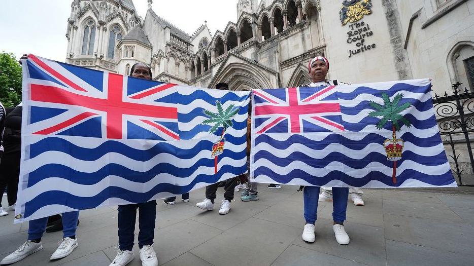 Two female protesters holding Chagos flags outside the High Court in central London, where a judge temporarily blocked on the government from concluding its negotiations