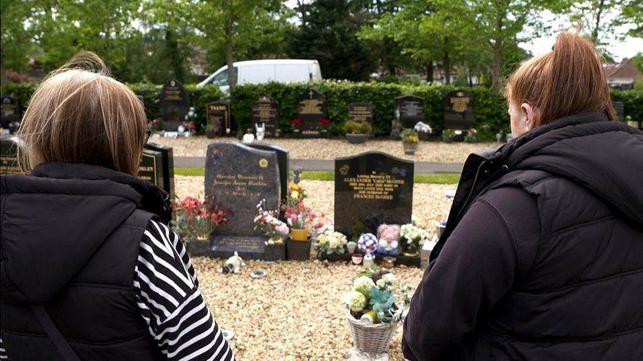 Two women stand with their backs to the camera, looking on at the grave of a loved one. Two gravestones stand before them, decorated with floral tributes. A row of stones sit behind that, then a hedge and a road behind it.