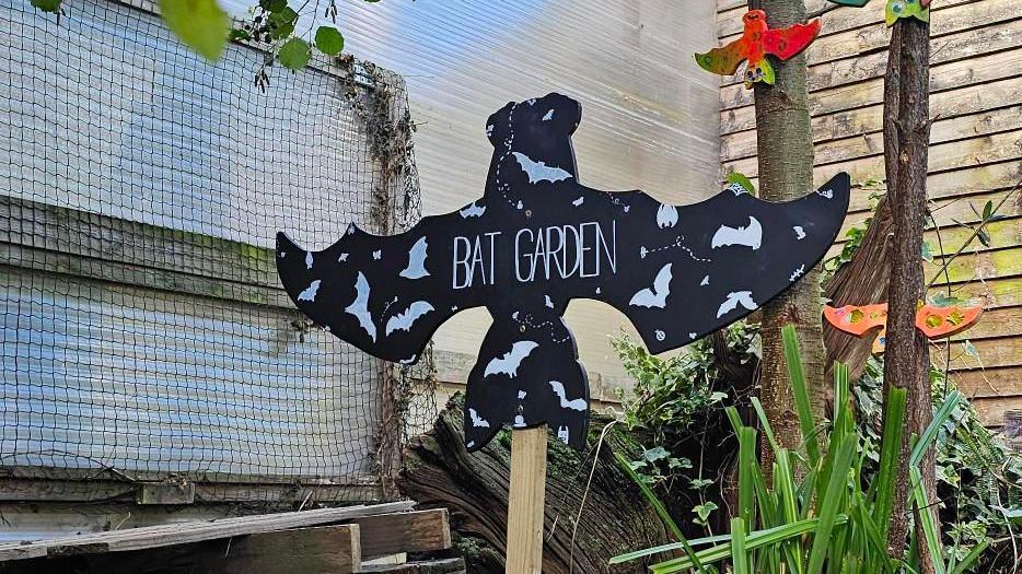 A sign is pictured within the garden. It is on a wooden stick and the sign is shaped like a bat. It reads "Bat Garden" in a large white font.