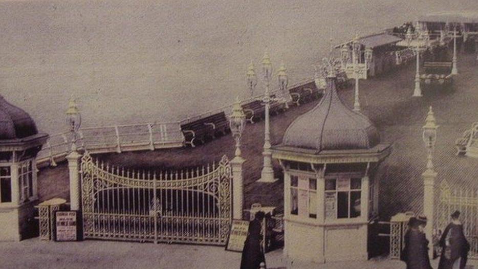 A black and white image of the old Cromer Pier. It has a gate and two small buildings either side of it at the entrance. On the pier are benches and lamp posts.