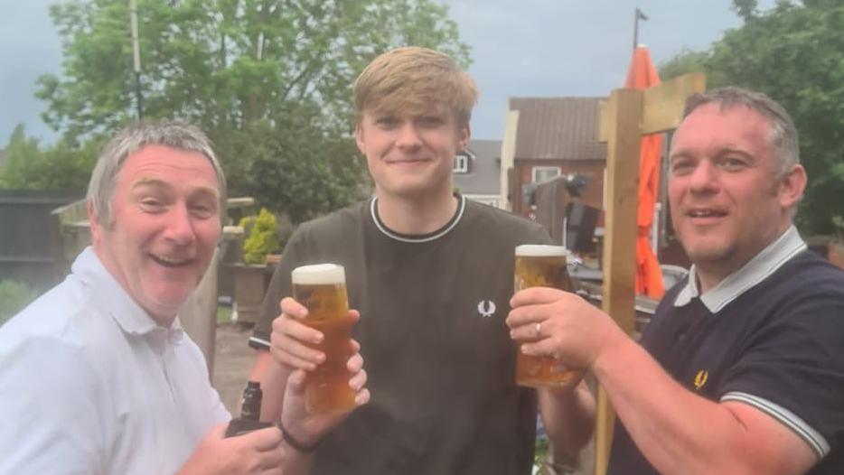 Three men in a line. The one in the middle is in his mid 20s, wearing a black top with white hems. Either side are two brothers in their fifties who look very similar, both with grey hair, wearing polo shirts and holding up pints.