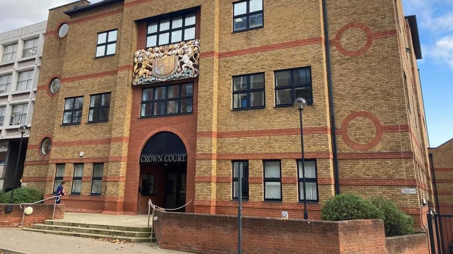 St Albans Crown Court: a brown-and-red brick building, with black-framed windows, in front of a low red-brick wall. The words "CROWN COURT" are written in gold capital letters above the entrance