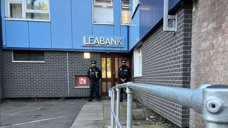 Two male police officers stand at the doors to the Leabank tower block of flats