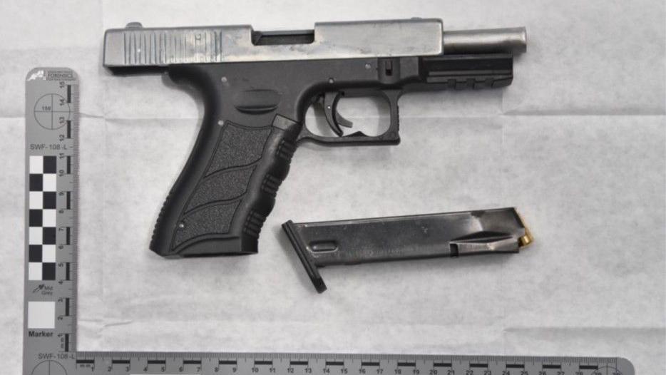 A disarmed gun lying flat on a table with the bullet chamber detached and lying beside it. It is a black handgun with a silver barrel. There is a measuring ruler laying next to it. 