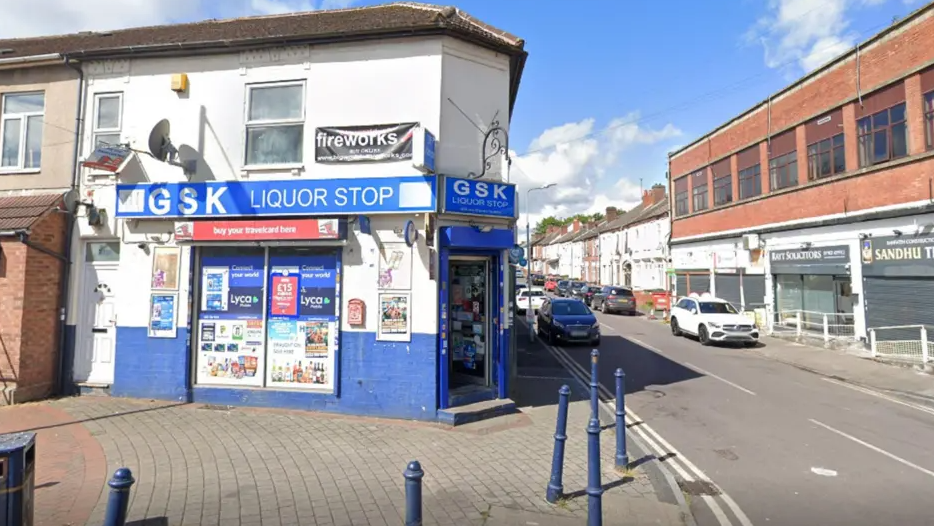 The shop has a blue and white frontage with its name above the door. It is on the corner of a street next to a taller brown brick building with a road separating them.