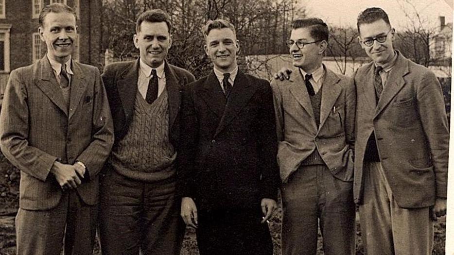 Fred Martin stands for a  photo with a group of four other young men. It is a black and white image and they are standing outdoors with an old house in the background, off to the side. Fred is looking sideways at the group and chuckling, while the others look at the camera smiling. Fred is wearing glasses and a suit, with his hair parted and slicked to one side. The other men wear suit jackets or winter coats, shirts and ties.