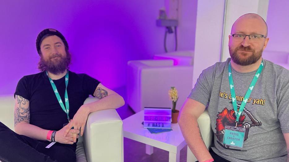 Two men - both bearded and wearing turquoise lanyards are sat in white armchairs in front of a purple-lit background. 