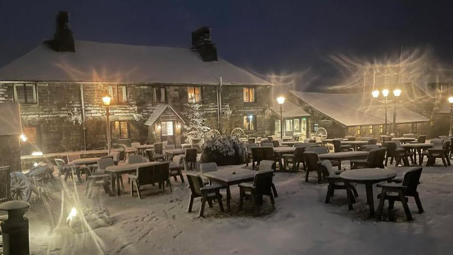 The outside of the Jamaica Inn covered in snow. Several tables and chairs are covered in a blanket of snow, as is the roof. It is dark.