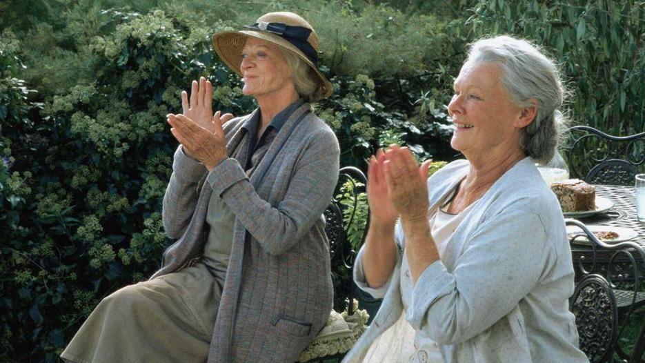 Dame Maggie Smith, left, and Dame Judi Dench, both wearing 1930s period dress, applaud and smile at something off camera - a table behind them has cake and drinks on it 
