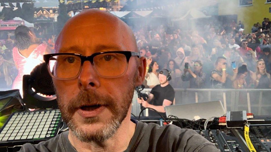 Nick Slater is looking at the camera with his back to a crowd at a gig or rave. He is balding with a beard and moustache and is wearing spectacles. Mixing decks can be seen on the stage behind him and there is a crowd of people dancing and filming on their phones as smoke effects surround them.