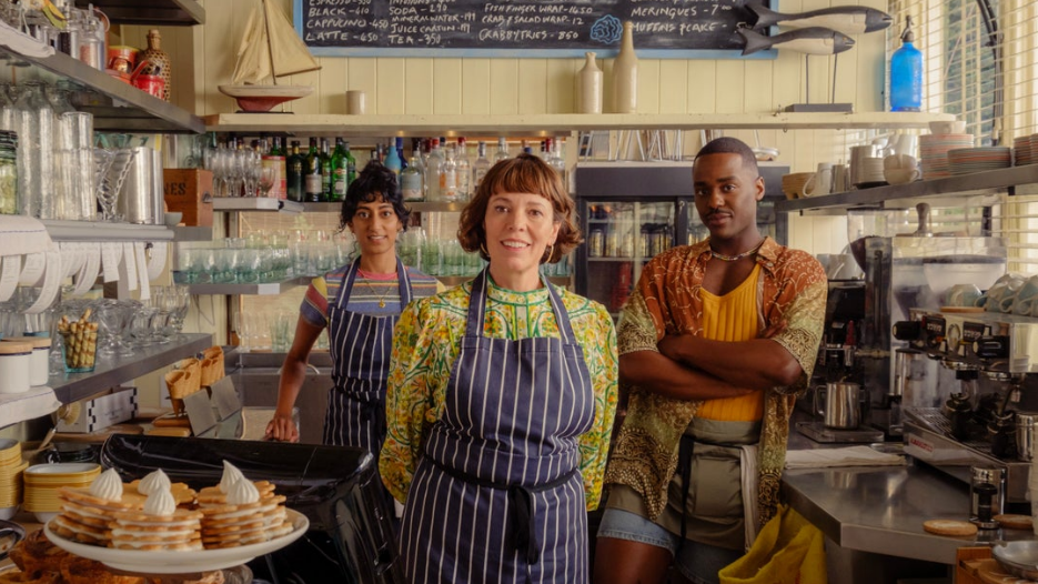 Three people stand behind a counter in a restaurant. They are wearing blue and white aprons. Mugs, glasses, alcohol and a coffee machine are in the background.