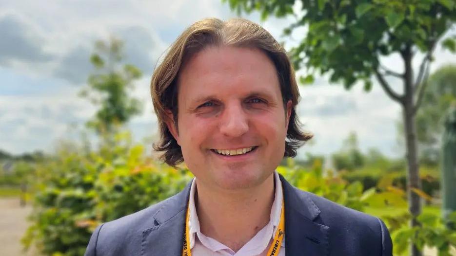 Liberal Democrat MP Steff Aquarone, a man with shoulder length, wearing a blue suit jacket and open necked shirt smiles for the camera on a sunny day