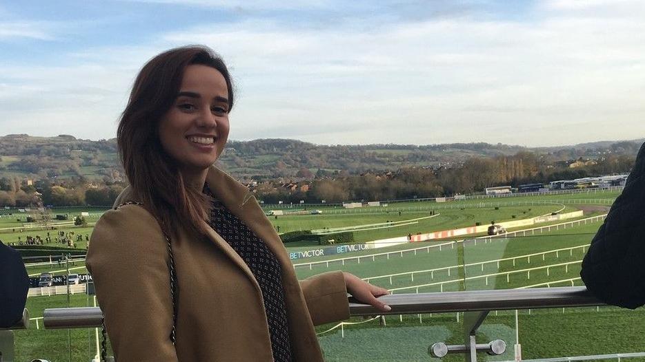 Ellie leaning on a railing, with a horse racecourse behind her.