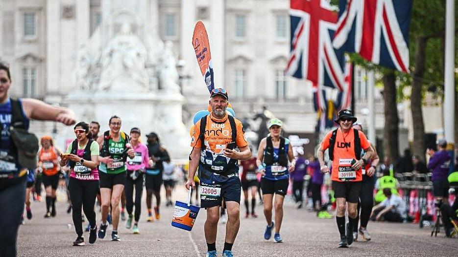 Rob Tiffney in all orange shirt walking the London Marathon whilst wearing a flag and carrying a donation bucket