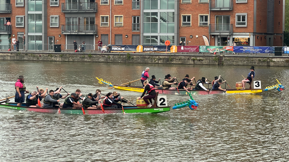 Two boats with 10 paddlers each are on the water in a city's docks. They are visibly straining to paddle hard to keep pace with each other