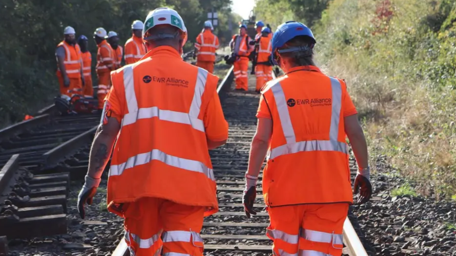 Railway engineers make their way along a stretch of track. They are wearing orange fluorescent jackets and trousers