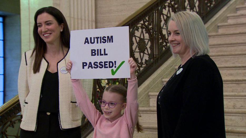 Kerry Boyd from Austisim NI and the DUP's Pam Cameron stand with a young girl holding a sign saying "Austism Bill passed" in 2022