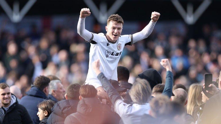 Jude Arthurs on shoulders on fans after promotion, both arms raised as fists abgove his head, smiling wearing Bromley home white kit