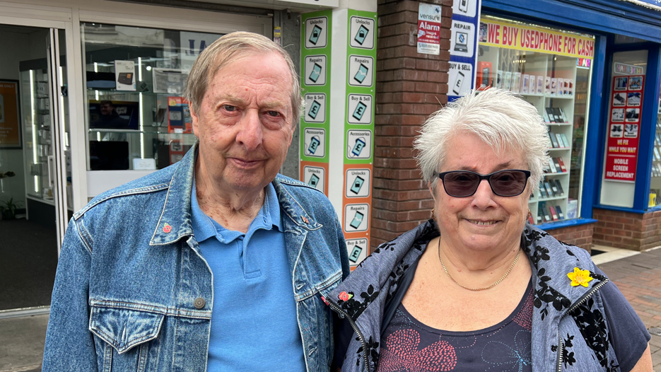 Brian Bosley wearing a jean jacket next to his wife standing in front of a phone shop