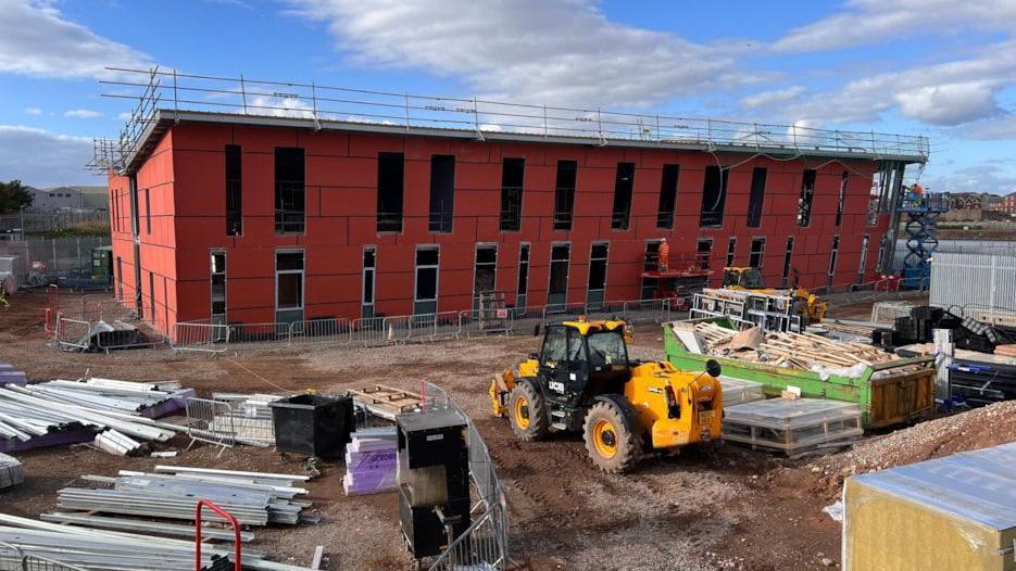 The building site of the University of Cumbria campus in Barrow. The building has no windows nor doors and is covered by red external panels. Building materials, a skip and a digger are in front of the building.