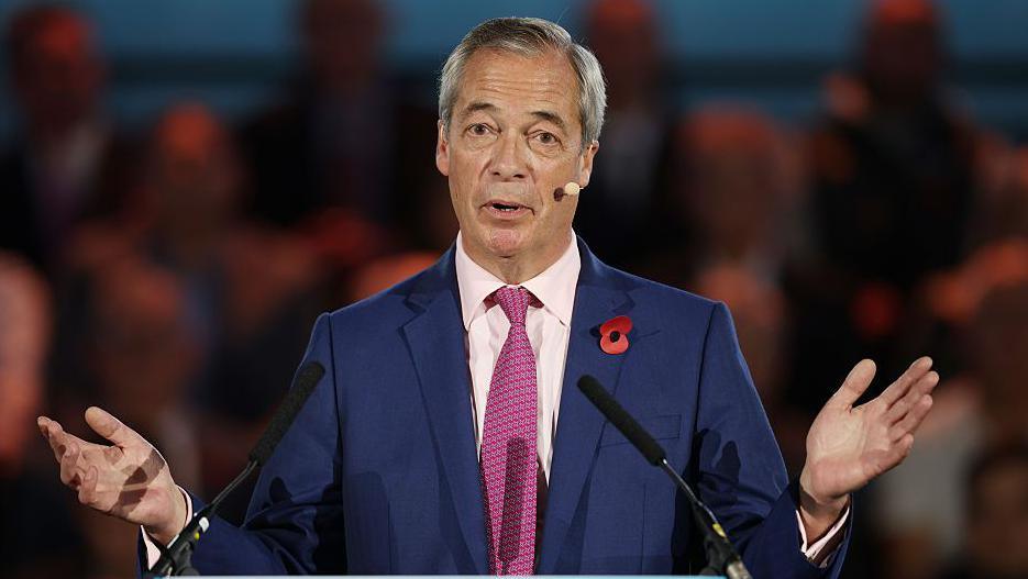 Nigel Farage wears a navy suit and a red tie. He has grey hair and has his arms out and palms open.