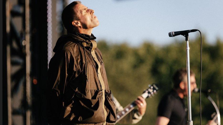 Liam Gallagher has his eyes closed with his head facing the sky while on stage. He is wearing a brown parka coat. To his left the arm of a band member playing guitar and Noel Gallagher playing guitar and singing, both blurred, can also be seen.