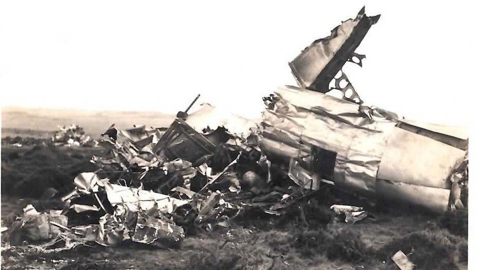 A black and white image of a plane crash. The plane is lying on grass in bits, with metal and wreckage strewn around