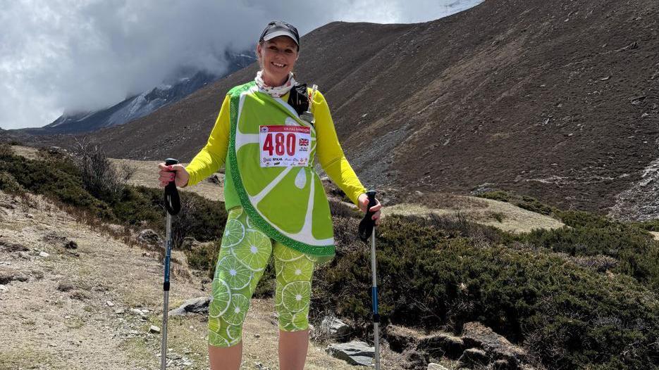 A woman who is dressed as a lime stands in front of a large mountain. She has leggings with pictures of the lime fruit on them plus a bright yellow long-sleeved shift and a top draped across her in green and white slices.