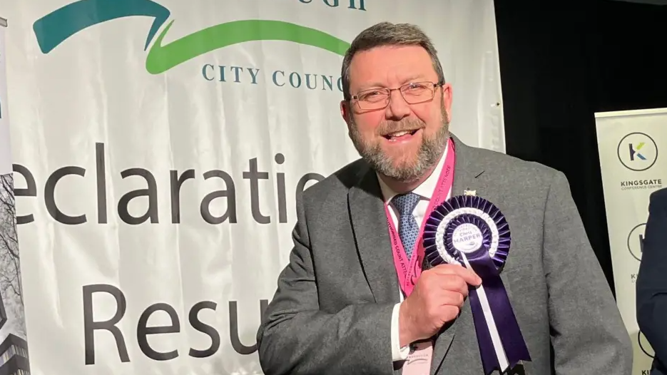 Chris Harper with short grey hair, a short beard and glasses. He is wearing a grey suit, white shirt and a blue tie, and is pointing to a round purple rosette pinned to his suit jacket following an election. He is smiling at the camera.