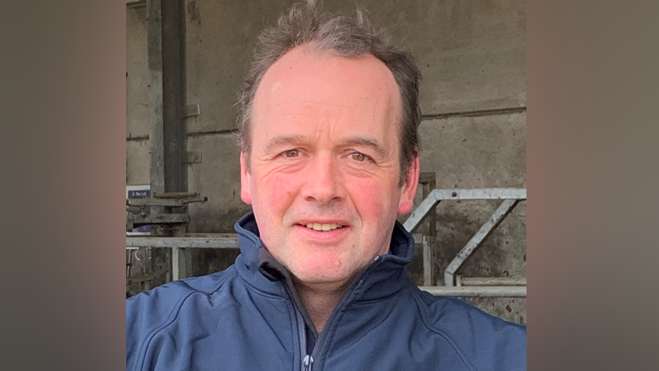 John Longmire is taking a selfie and smiling slightly at the camera. He has short brown hair and is wearing a blue zip-up jacket.