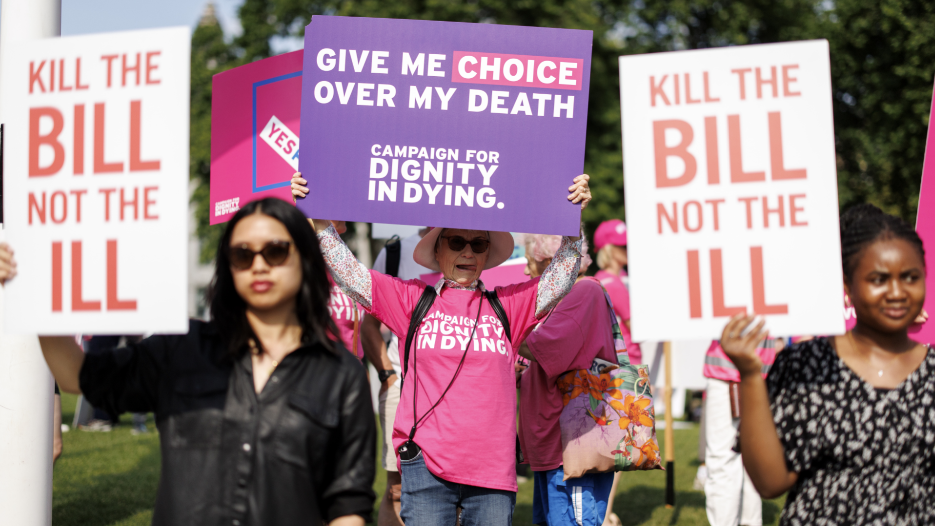 Campaigners outside Parliament hold up placards. Two women are holding a placard reading: "Kill the bill not the ill". Another woman has a poster which say: "Give me choice over my death."