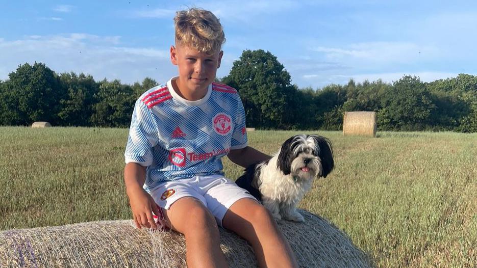 A young, blonde boy in a blue and white Manchester United kit sitting on top of a haybale with a small, black and white dog.