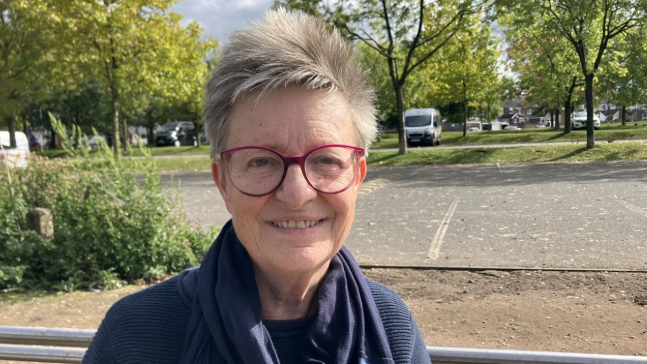 A woman with short grey hair swept upwards, wearing red rimmed glasses and a blue cowled top. She is standing outside, with trees behind her, and a number of vehicles parked up