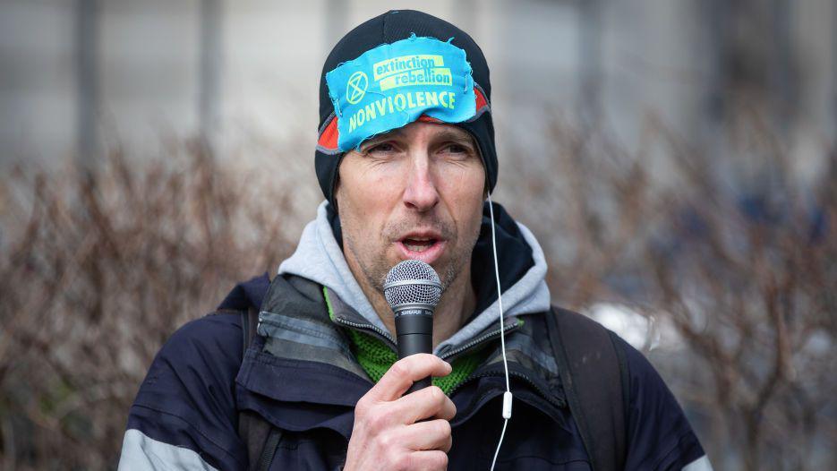 Etienne Stott holding a microphone and talking into it, while wearing a hat