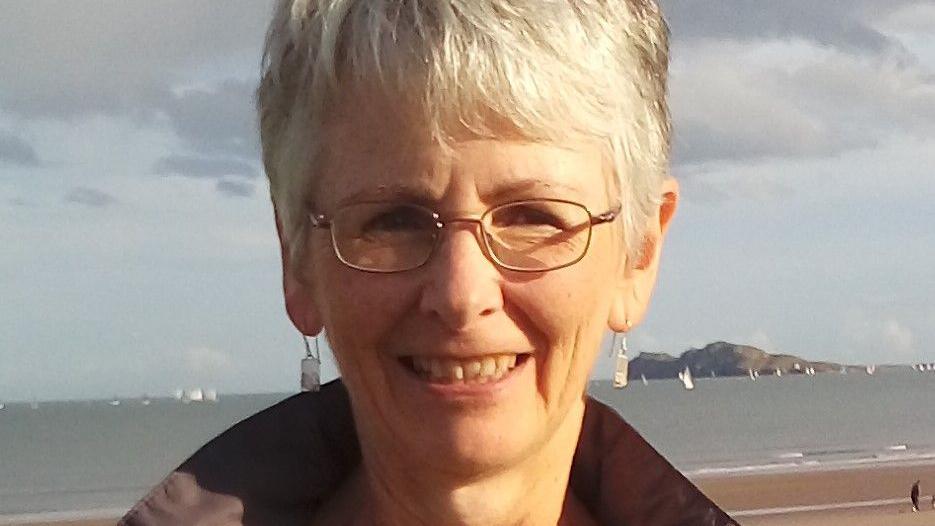 Pensioner Olwen Jones standing on a beach with sailing boats in the bay behind her, says she gives her WFP to charity