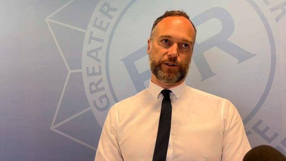 Detective Superintendent Joseph Harrop, wearing a white shirt with a dark blue tie, speaks to the camera against a backdrop showing the Greater Manchester Police badge