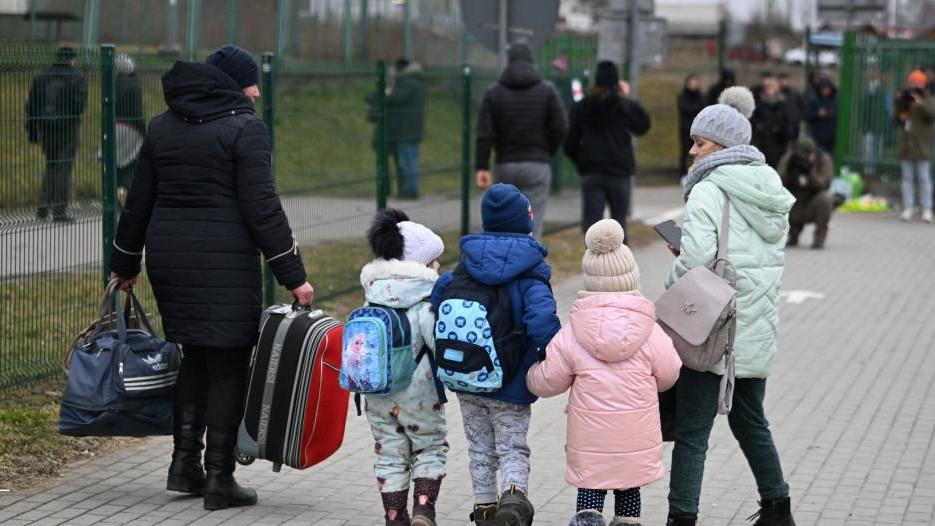 Three young children hold hands with their backs to the camera as a woman looks down at them and a man on the other side carries two bags