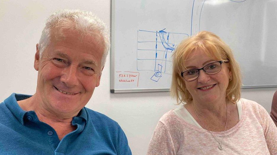 A man in a blue shirt sits next to a woman in a pink top with glasses and both are smiling and looking happy.