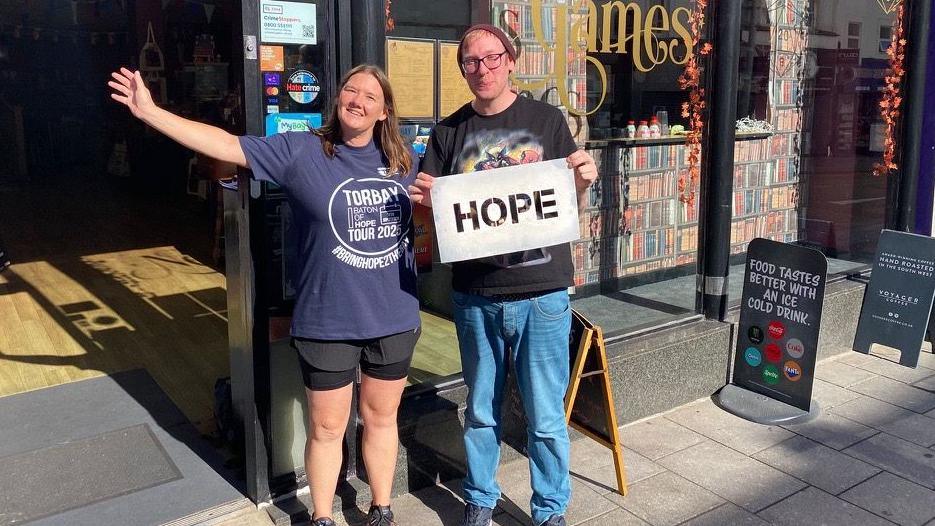 The image shows two people standing outside a shop called Nexus Games, which has large front windows and gold lettering. One person is holding a sign that says "HOPE", while the other wears a blue T-shirt.